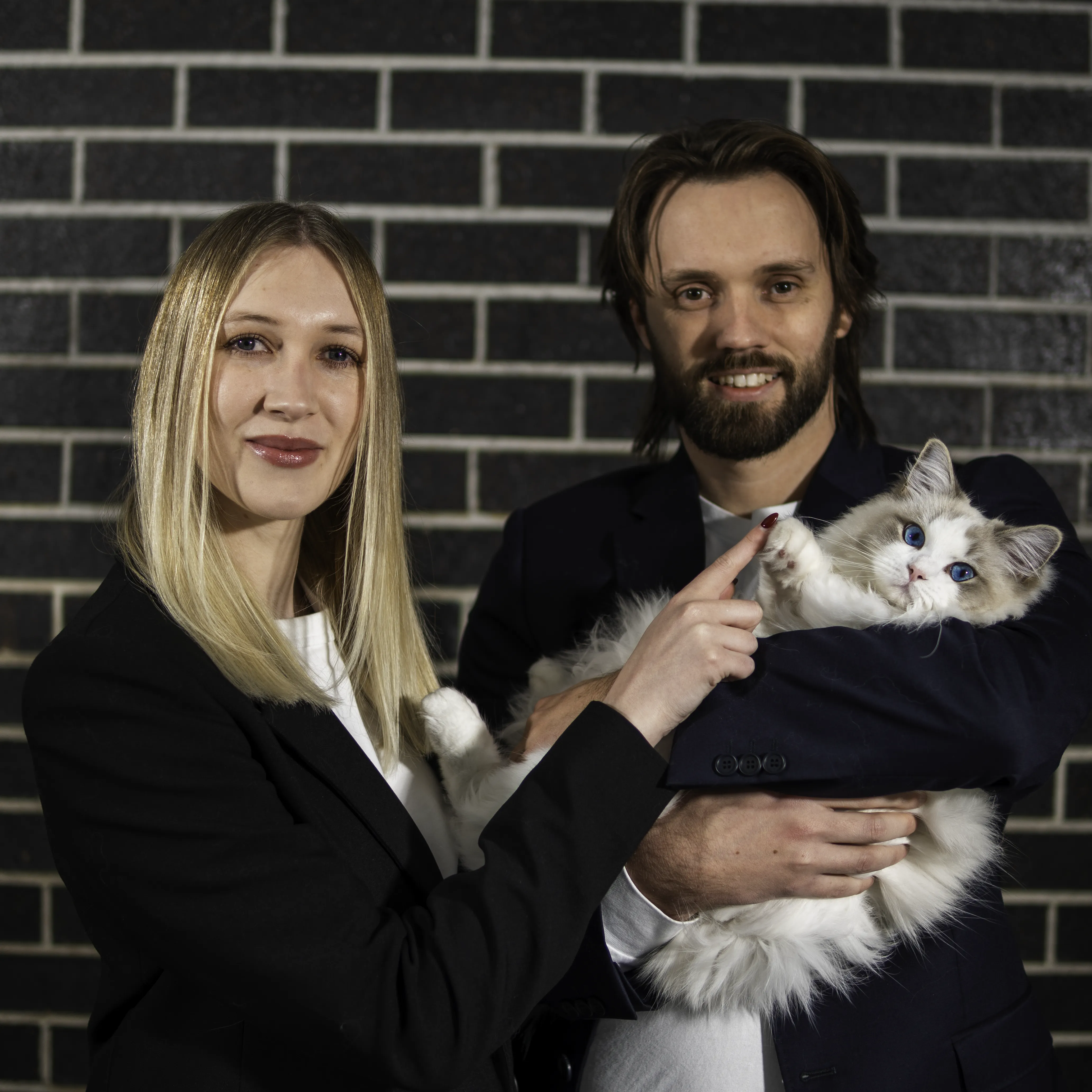 Brie and Jared, co-founders of Maeve Social, standing against a dark brick wall holding their ragdoll cat